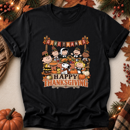 A black short-sleeve graphic T-shirt laid flat on a neutral surface with autumn props. The shirt features a large Thanksgiving-themed cartoon print with Peanuts characters, pumpkins, and the words 'GIVE THANKS' and 'HAPPY THANKSGIVING.