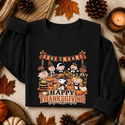 A black long-sleeve sweatshirt laid flat on a cozy autumn-themed background. The sweatshirt features a colorful graphic of Peanuts characters celebrating Thanksgiving, with pumpkins, a turkey, and the text 'GIVE THANKS' and 'HAPPY THANKSGIVING'.