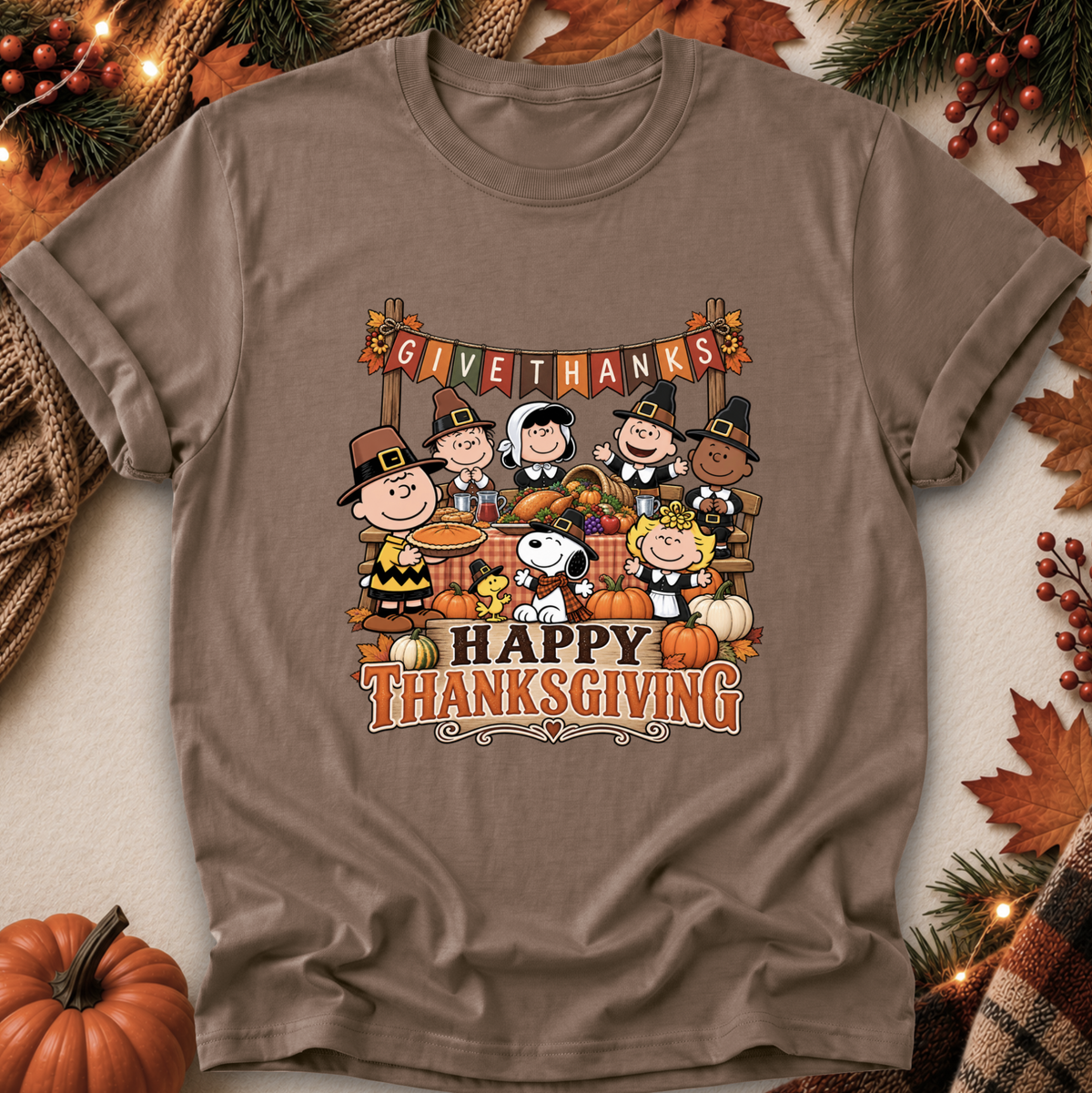 A brown short-sleeve crewneck t-shirt featuring a colorful cartoon graphic of Peanuts characters celebrating Thanksgiving, surrounded by autumn leaves, pumpkins, and pine branches.