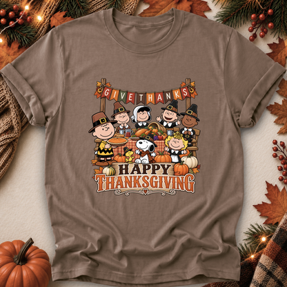 A brown short-sleeve crewneck t-shirt featuring a colorful cartoon graphic of Peanuts characters celebrating Thanksgiving, surrounded by autumn leaves, pumpkins, and pine branches.