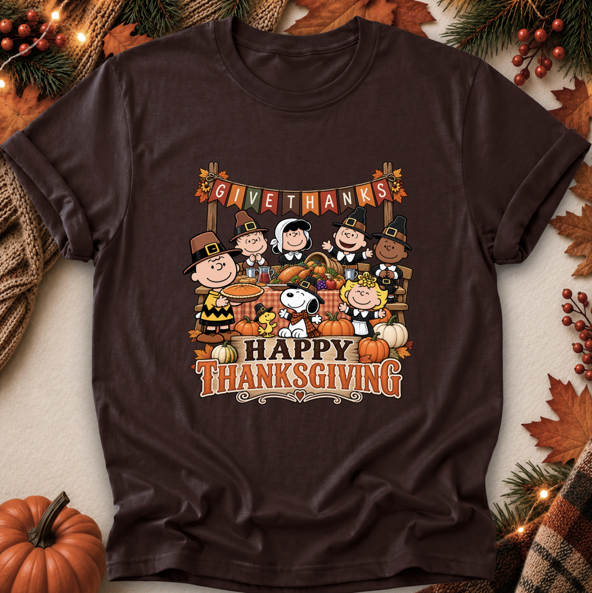 A dark brown short-sleeve t-shirt featuring a colorful cartoon graphic of Peanuts characters celebrating Thanksgiving, with 'GIVE THANKS' and 'HAPPY THANKSGIVING' text. The shirt is displayed in a flat-lay setting surrounded by autumn-themed props including pumpkins, pine branches, and leaves.