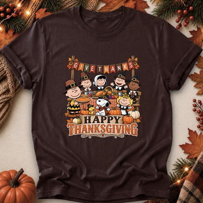 A dark brown short-sleeve t-shirt featuring a colorful cartoon graphic of Peanuts characters celebrating Thanksgiving, with 'GIVE THANKS' and 'HAPPY THANKSGIVING' text. The shirt is displayed in a flat-lay setting surrounded by autumn-themed props including pumpkins, pine branches, and leaves.