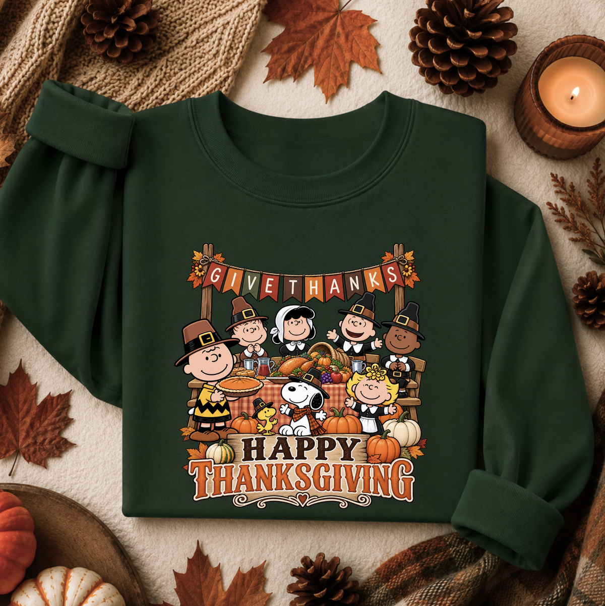 A dark green long-sleeve sweatshirt with a colorful Thanksgiving graphic featuring Peanuts characters, pumpkins, and the text 'GIVE THANKS Happy THANKSGIVING', styled in a flat lay with autumn leaves, pinecones, a candle, and knitted fabric.