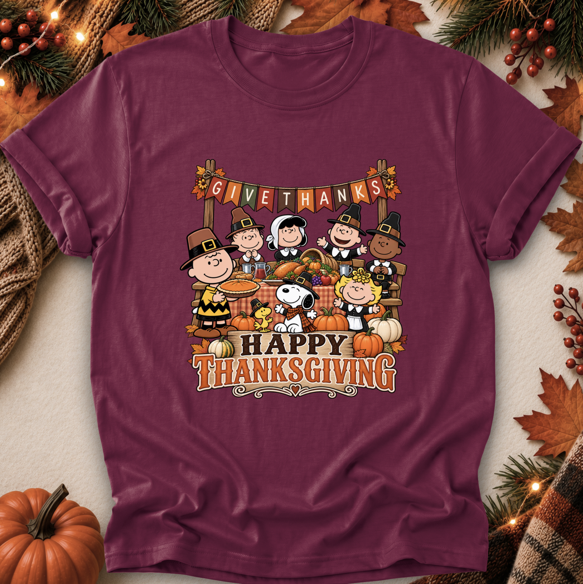 A maroon short-sleeve graphic T-shirt laid flat with autumn props around it. The front print shows Peanuts characters gathered around pumpkins with a banner reading 'GIVE THANKS' and large text reading 'HAPPY THANKSGIVING.
