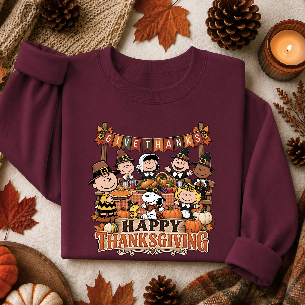 A maroon long-sleeve sweatshirt laid flat on a neutral fabric background, surrounded by autumn props including pinecones, leaves, a candle, and small pumpkins. The sweatshirt features a large Thanksgiving graphic with Peanuts characters and the text 'GIVE THANKS HAPPY THANKSGIVING'.