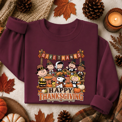 A maroon long-sleeve sweatshirt laid flat on a neutral fabric background, surrounded by autumn props including pinecones, leaves, a candle, and small pumpkins. The sweatshirt features a large Thanksgiving graphic with Peanuts characters and the text 'GIVE THANKS HAPPY THANKSGIVING'.