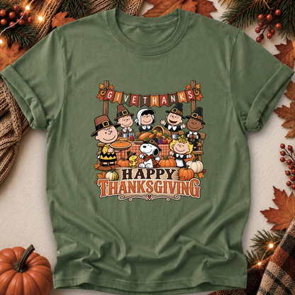 An olive green short-sleeve t-shirt featuring a colorful 'Happy Thanksgiving' graphic with Peanuts characters, styled in a flat-lay with autumn props like pumpkins, pine branches, and leaves.