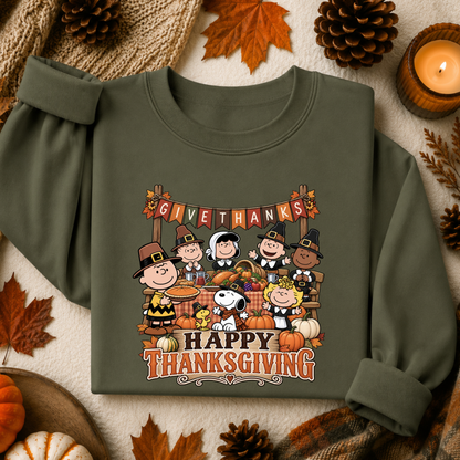 An olive green long-sleeve sweatshirt with a colorful 'Happy Thanksgiving' graphic featuring Peanuts characters, styled in a flat-lay with autumn leaves, pinecones, and a candle.