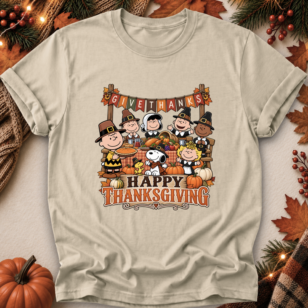 A light beige graphic t-shirt featuring the Peanuts characters Snoopy, Woodstock, Charlie Brown, and others in Thanksgiving attire with pumpkins and autumn decorations. The shirt is displayed in a flat-lay arrangement with seasonal props like pine branches, leaves, and a small pumpkin.