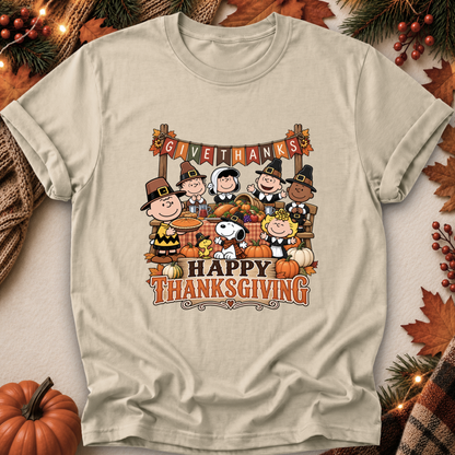 A light beige graphic t-shirt featuring the Peanuts characters Snoopy, Woodstock, Charlie Brown, and others in Thanksgiving attire with pumpkins and autumn decorations. The shirt is displayed in a flat-lay arrangement with seasonal props like pine branches, leaves, and a small pumpkin.