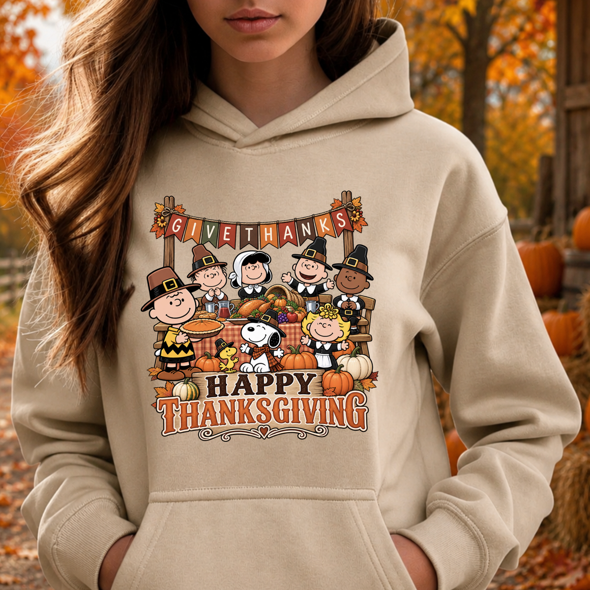A woman wearing a beige hoodie with a colorful Peanuts Thanksgiving graphic on the front, set against an autumn outdoor background with trees and pumpkins.