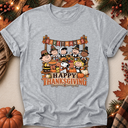 A heather gray short-sleeve t-shirt featuring a colorful graphic of Peanuts characters celebrating Thanksgiving, surrounded by autumn leaves, pine branches, and a pumpkin in a styled flat-lay setting.