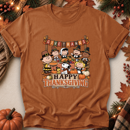 A rust-orange short-sleeve t-shirt featuring a colorful 'GIVE THANKS Happy Thanksgiving' Peanuts graphic, styled in a flat-lay with autumn decorations like pumpkins, pine branches, and a plaid scarf.