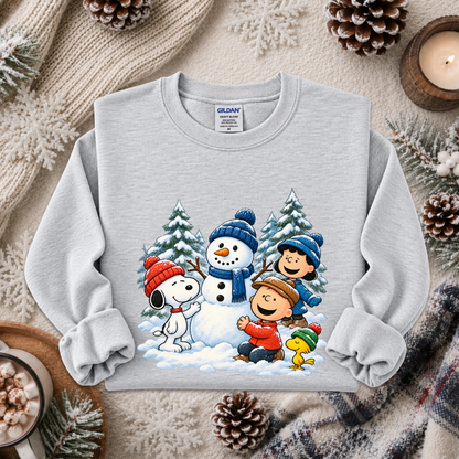 A light gray crewneck sweatshirt featuring a colorful graphic of Peanuts characters Snoopy, Woodstock, Charlie Brown, and Pepple in a winter scene, styled on a cozy holiday flat-lay with a knit blanket, pinecones, and a candle.