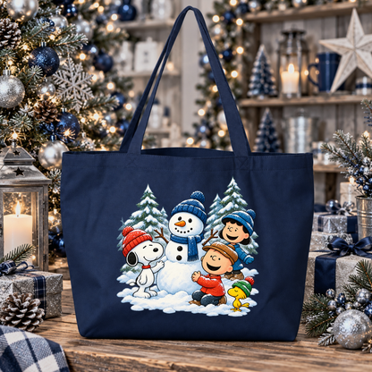A navy blue canvas tote bag featuring a festive Christmas print of Snoopy, Charlie Brown, Woodstock, and a snowman in winter clothing, displayed on a wooden table in front of a decorated Christmas tree and holiday ornaments.