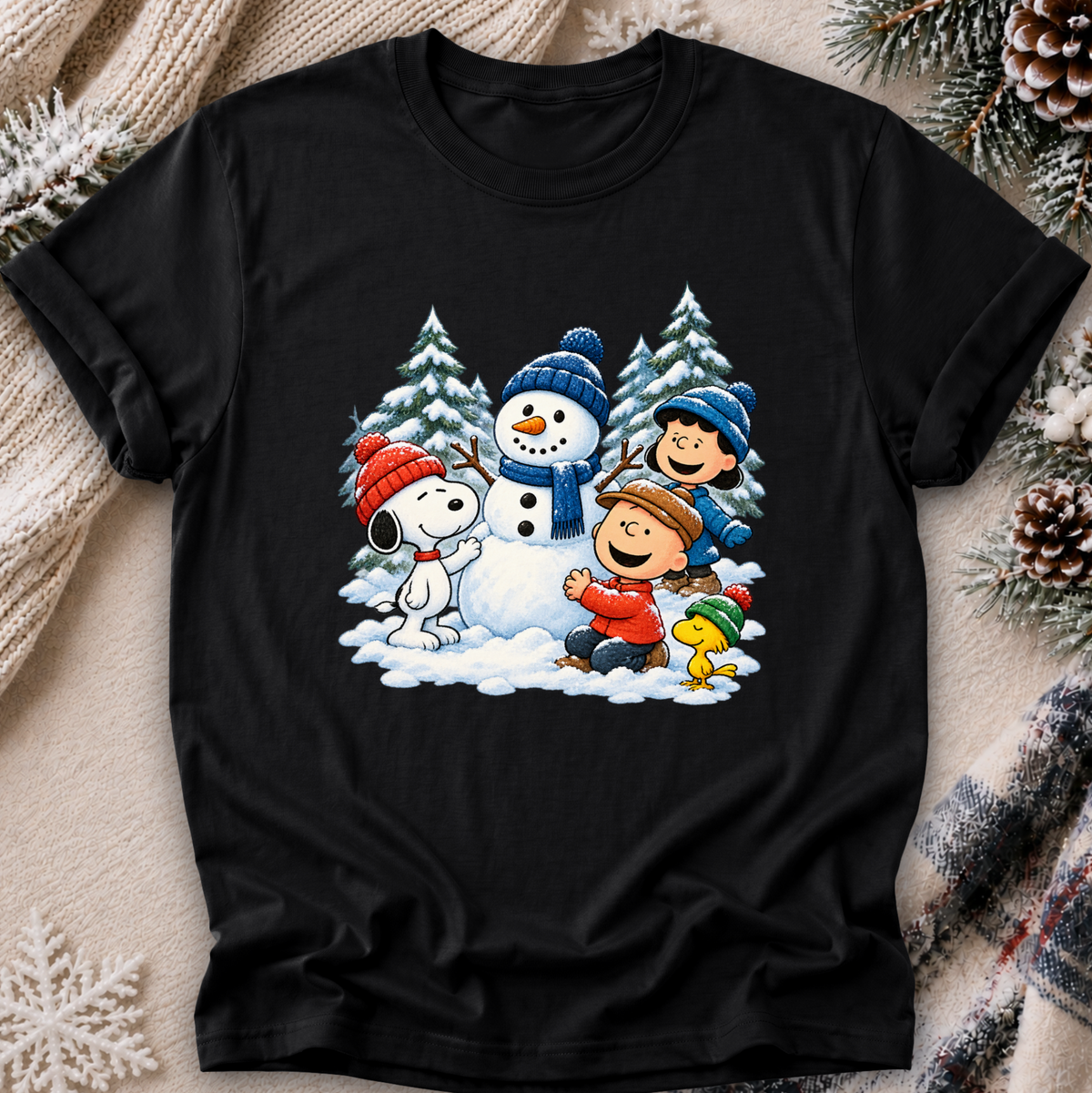A black short-sleeve crewneck t-shirt featuring a colorful graphic print of Peanuts characters Snoopy, Woodstock, Charlie Brown, and Peanuts in a winter snow scene. The t-shirt is displayed flat on a cozy background with a beige knit sweater, pinecones, and a plaid scarf.