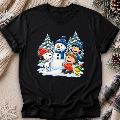 A black short-sleeve crewneck t-shirt featuring a colorful graphic print of Peanuts characters Snoopy, Woodstock, Charlie Brown, and Peanuts in a winter snow scene. The t-shirt is displayed flat on a cozy background with a beige knit sweater, pinecones, and a plaid scarf.