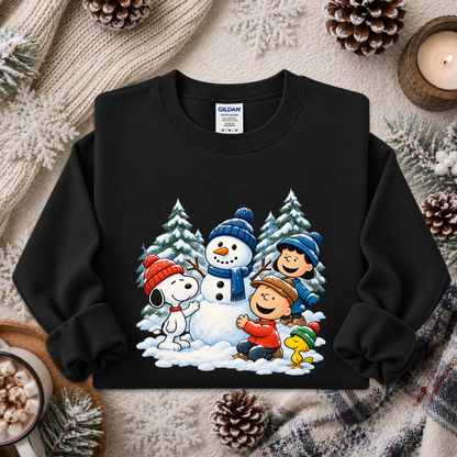 A black crewneck sweatshirt featuring a colorful graphic of Peanuts characters Snoopy, Woodstock, Charlie Brown, and Bob Brown in a snowy winter scene. The sweatshirt is displayed in a flat-lay setting on a cozy beige knit blanket, surrounded by pinecones, a lit candle, and a mug of hot cocoa.