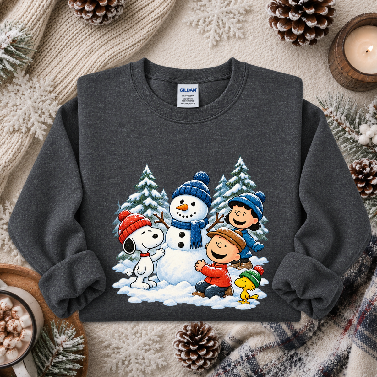A dark gray crewneck sweatshirt featuring a colorful graphic of Peanuts characters Snoopy, Woodstock, Charlie Brown, and Lucy in a winter scene, styled on a cozy holiday background with a knit blanket, pinecones, and a candle.