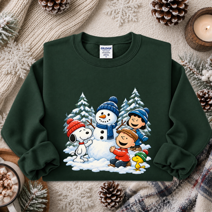 A dark green crewneck sweatshirt featuring a colorful graphic of Peanuts characters Snoopy, Woodstock, Charlie Brown, Peanuts, and the snowman in a winter scene. The sweatshirt is styled in a cozy flat-lay arrangement with pinecones, a candle, and a knit blanket.