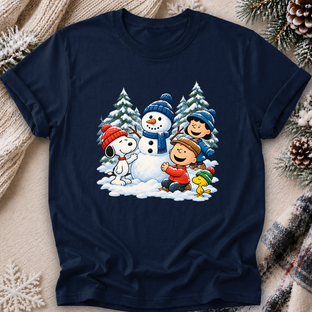 A navy blue tshirt featuring a colorful graphic of Peanuts characters Snoopy, Woodstock, Charlie Brown, and Lucy building a snowman in the snow. The sweatshirt is styled in a cozy flat-lay setting with a knitted beige scarf, pinecones, and a lit candle.
