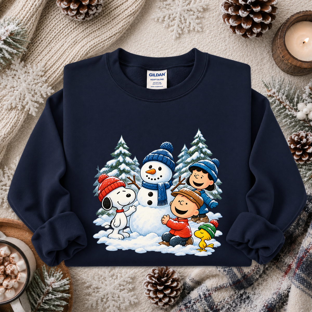 A navy blue crewneck sweatshirt featuring a colorful graphic of Peanuts characters Snoopy, Woodstock, Charlie Brown, and Lucy building a snowman in the snow. The sweatshirt is styled in a cozy flat-lay setting with a knitted beige scarf, pinecones, and a lit candle.