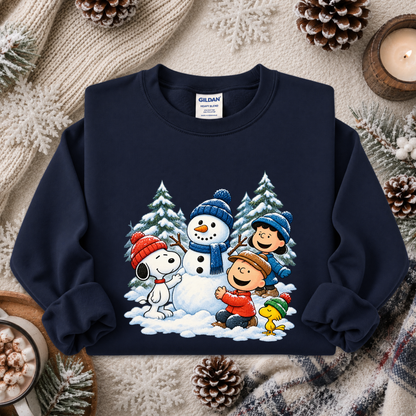 A navy blue crewneck sweatshirt featuring a colorful graphic of Peanuts characters Snoopy, Woodstock, Charlie Brown, and Lucy building a snowman in the snow. The sweatshirt is styled in a cozy flat-lay setting with a knitted beige scarf, pinecones, and a lit candle.