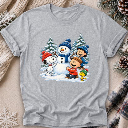 A gray tshirt featuring a colorful graphic of Peanuts characters Snoopy, Woodstock, Charlie Brown, and Lucy building a snowman in the snow. The sweatshirt is styled in a cozy flat-lay setting with a knitted beige scarf, pinecones, and a lit candle.
