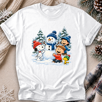 A white short-sleeve crewneck t-shirt featuring a colorful cartoon graphic of Peanuts characters Snoopy, Woodstock, Charlie Brown, and Linus building a snowman in the snow. The shirt is displayed flat on a cozy background with a beige knit sweater, pinecones, and a plaid scarf.