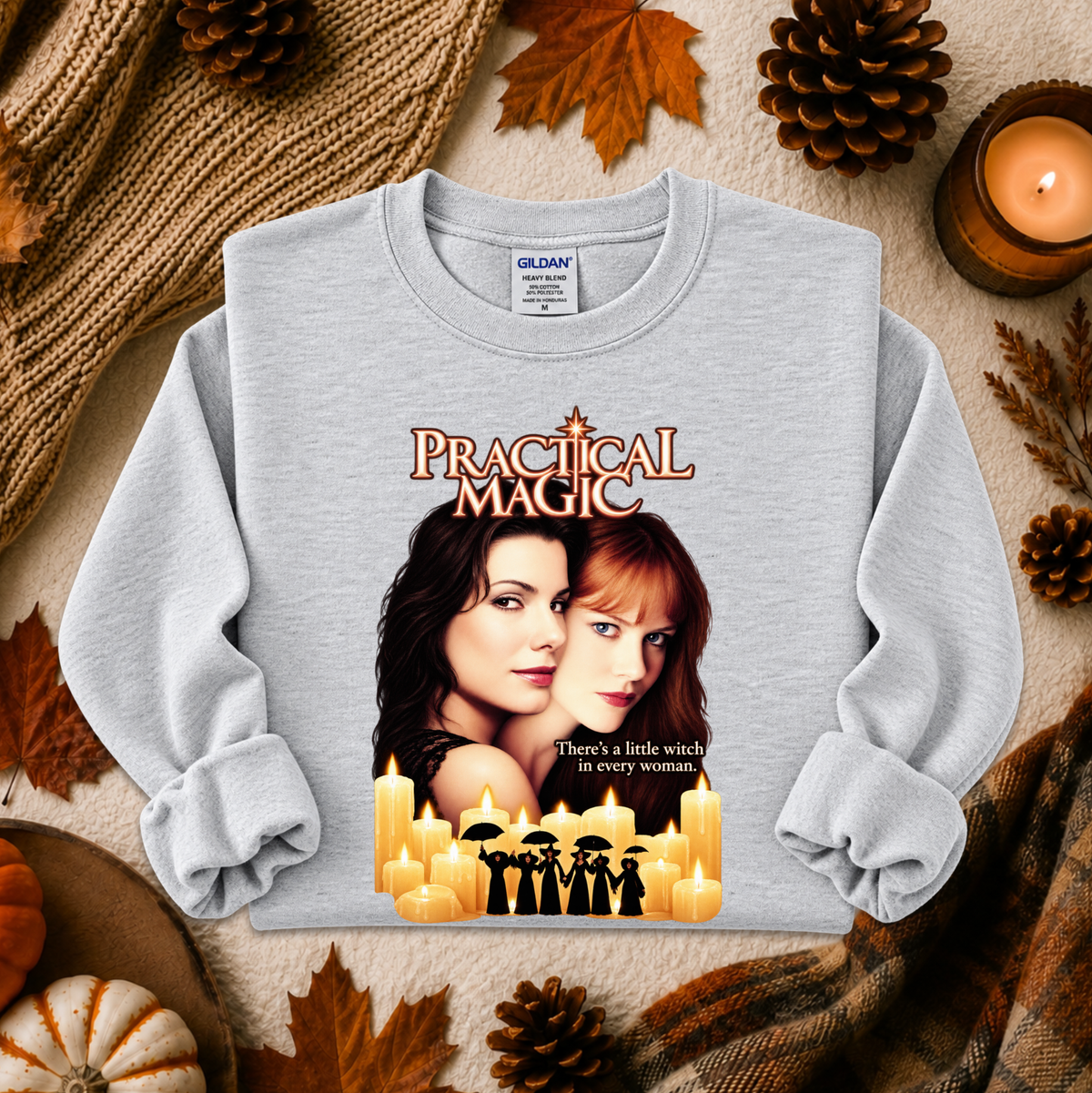 A light gray crewneck sweatshirt featuring a 'Practical Magic' movie graphic print, styled in a cozy flat-lay arrangement with autumn leaves, pinecones, a knitted scarf, and a lit candle. The Gildan brand tag is visible inside the collar.