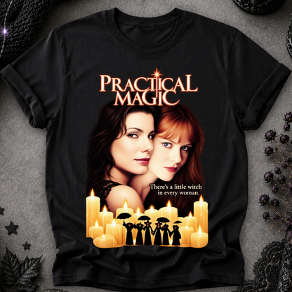 A black short-sleeve graphic T-shirt laid flat on a textured gray surface with decorative dark lace and dried flowers around it. The shirt features a large printed design from the movie 'Practical Magic,' including the title in orange-gold lettering, two women's faces, glowing candles, and the quote 'There's a little witch in every woman.
