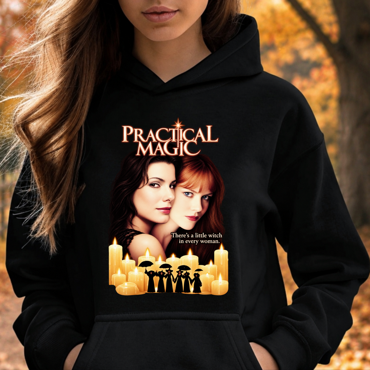 A woman wearing a black hoodie with a 'Practical Magic' graphic print featuring the movie title, two women's faces, candles, and silhouettes of witches, set against an autumn outdoor background.