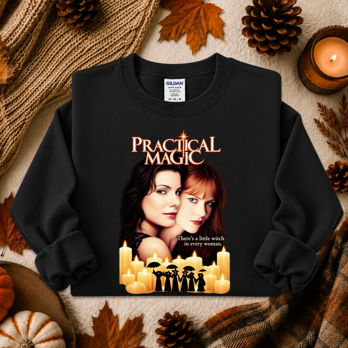 A black long-sleeve crewneck sweatshirt featuring a 'Practical Magic' movie graphic print, styled in a flat-lay with autumn props like pinecones, leaves, and a candle.