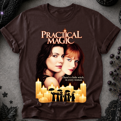 A dark brown graphic t-shirt featuring the movie 'Practical Magic' design with images of AnnaSophia Roback and Nicole Kidman, surrounded by glowing candles and the tagline 'There's a little witch in every woman.' The shirt is displayed flat on a textured gray background with gothic decorative props.