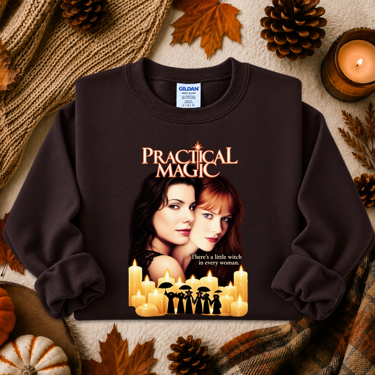 A dark brown crewneck sweatshirt featuring a 'Practical Magic' movie graphic print, styled in an autumn flat-lay setting with pinecones, leaves, and a candle.