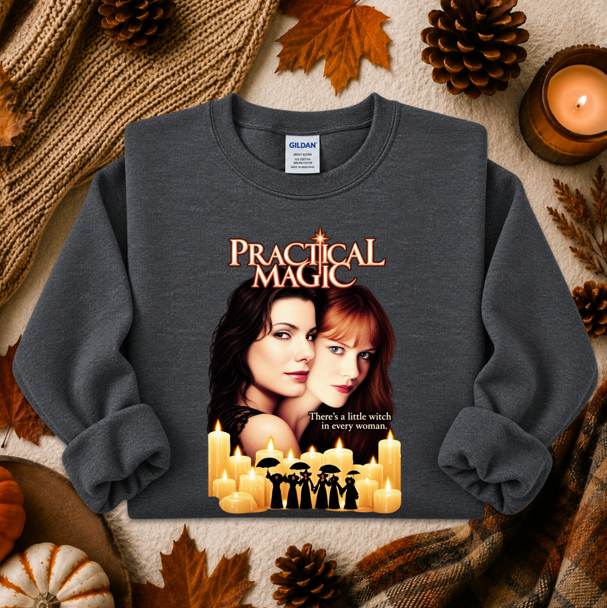 A dark gray crewneck sweatshirt featuring a 'Practical Magic' movie graphic, styled in a cozy autumn flat-lay with pinecones, leaves, a candle, and a knitted blanket.