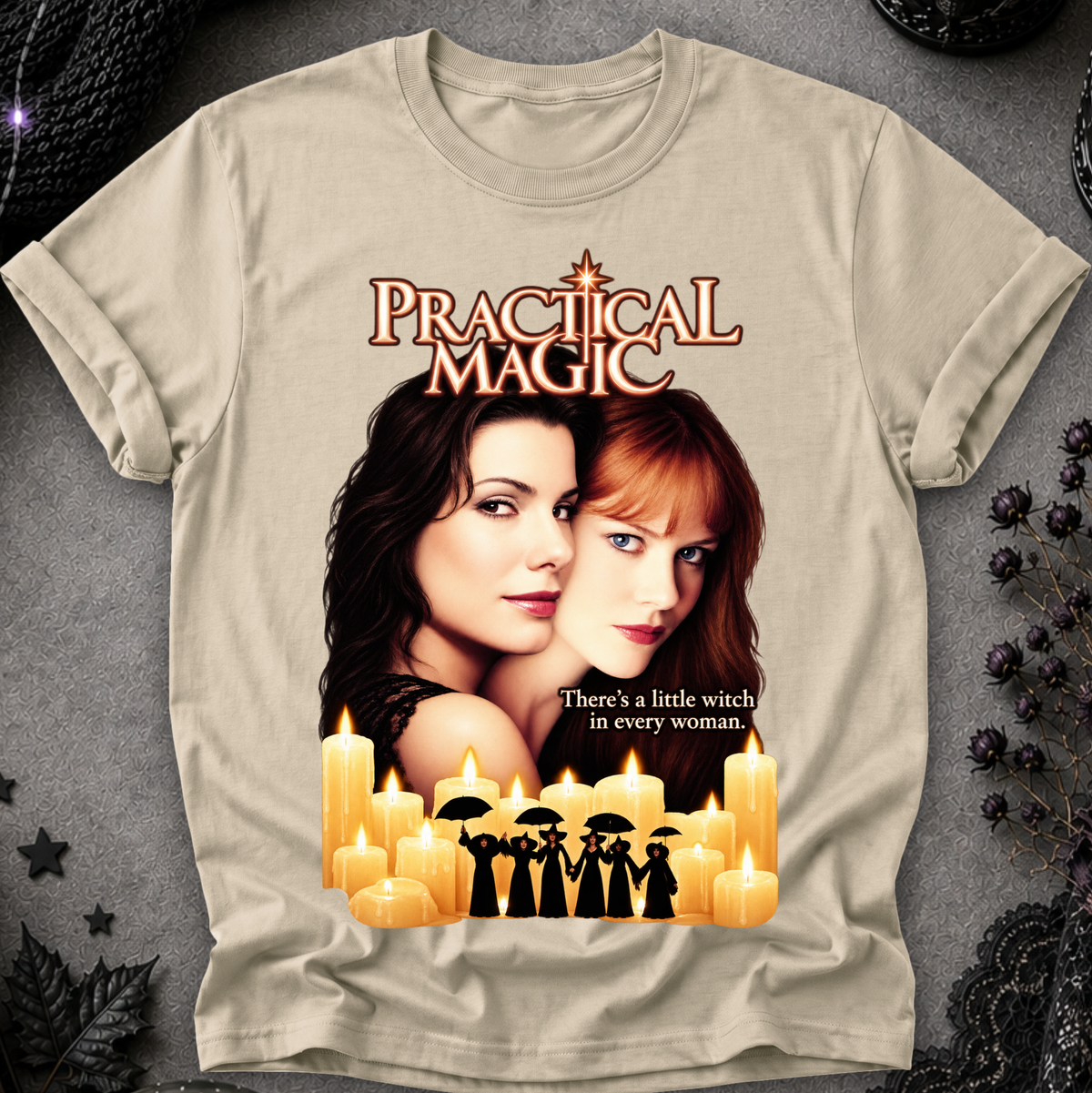 A beige graphic t-shirt featuring a 'Practical Magic' movie print with Anne Hathaway and Jennifer Garner, surrounded by candles and the text 'There's a little witch in every woman.' The shirt is displayed flat on a dark gray textured background with gothic decorative props.