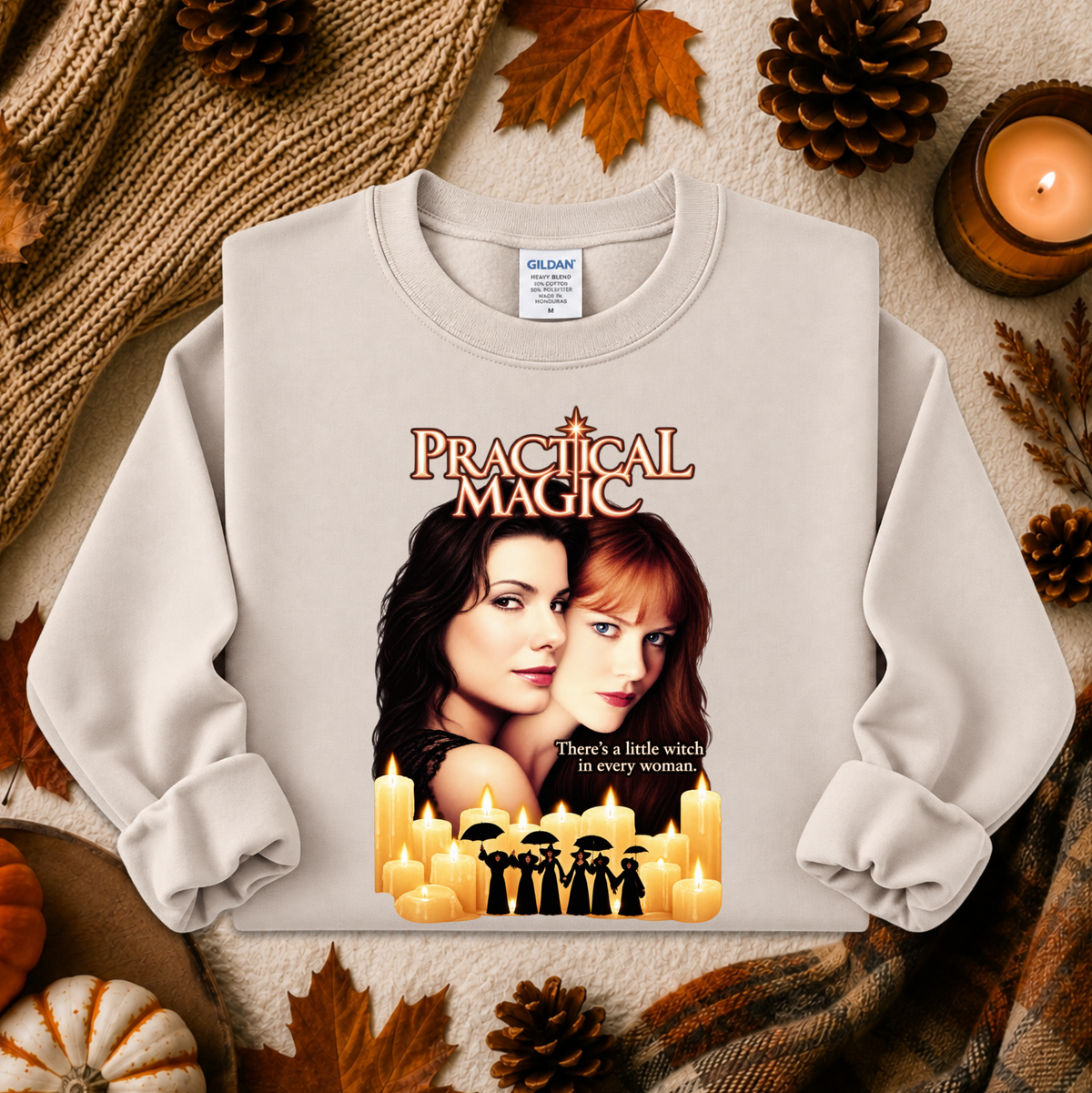A light beige crewneck sweatshirt featuring a 'Practical Magic' graphic print with two women and candles, styled in an autumn flat-lay with pinecones, leaves, and a candle.