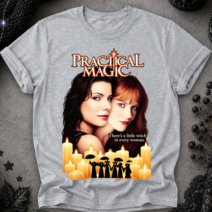 A heather gray graphic t-shirt featuring a 'Practical Magic' movie design with a portrait of two women, numerous lit candles, and the tagline 'There's a little witch in every woman.' The shirt is displayed flat on a textured gray background decorated with gothic props like dried flowers and lace.
