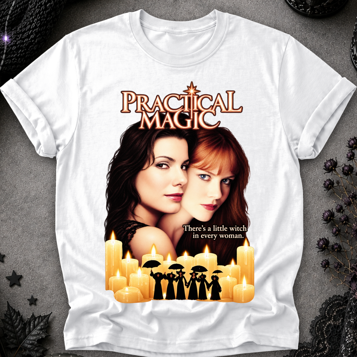 A white short-sleeve graphic T-shirt laid flat on a dark textured surface. The shirt features a large printed design with the words 'Practical Magic,' two women's portraits, glowing candles, and small witch silhouettes with umbrellas.