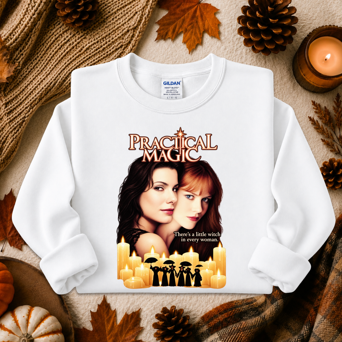 A white long-sleeve crewneck sweatshirt featuring a 'Practical Magic' movie graphic print, styled in a cozy autumn flat-lay with pinecones, leaves, and candles. The Gildan brand label is visible inside the collar.