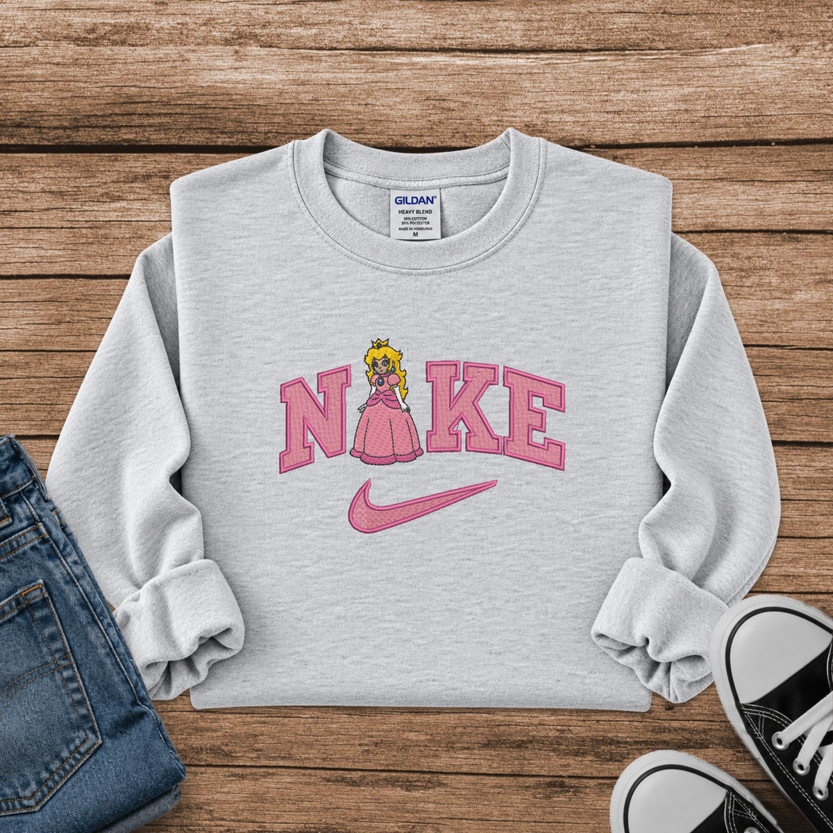 A light heather gray sweatshirt laid flat on a rustic wood background, featuring a pink graphic that reads 'N' and 'KE' with a cartoon princess peach in the middle and a pink Nike swoosh underneath. Partial jeans and black sneakers are visible at the edges as styling props.