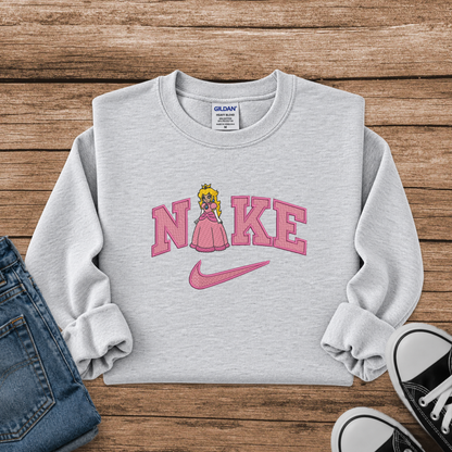 A light heather gray sweatshirt laid flat on a rustic wood background, featuring a pink graphic that reads 'N' and 'KE' with a cartoon princess peach in the middle and a pink Nike swoosh underneath. Partial jeans and black sneakers are visible at the edges as styling props.