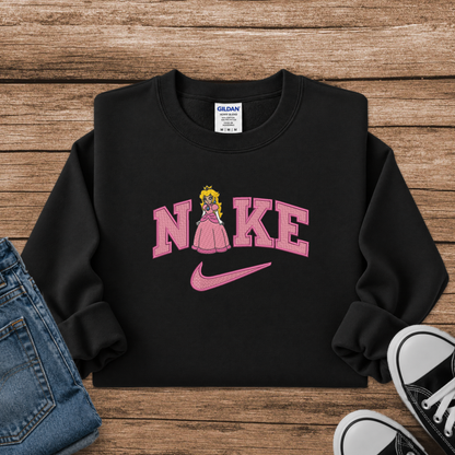 A black crewneck sweatshirt with a pink 'Nike' graphic featuring a princess character and a pink swoosh, styled in a flat lay on a wooden background with jeans and sneakers.