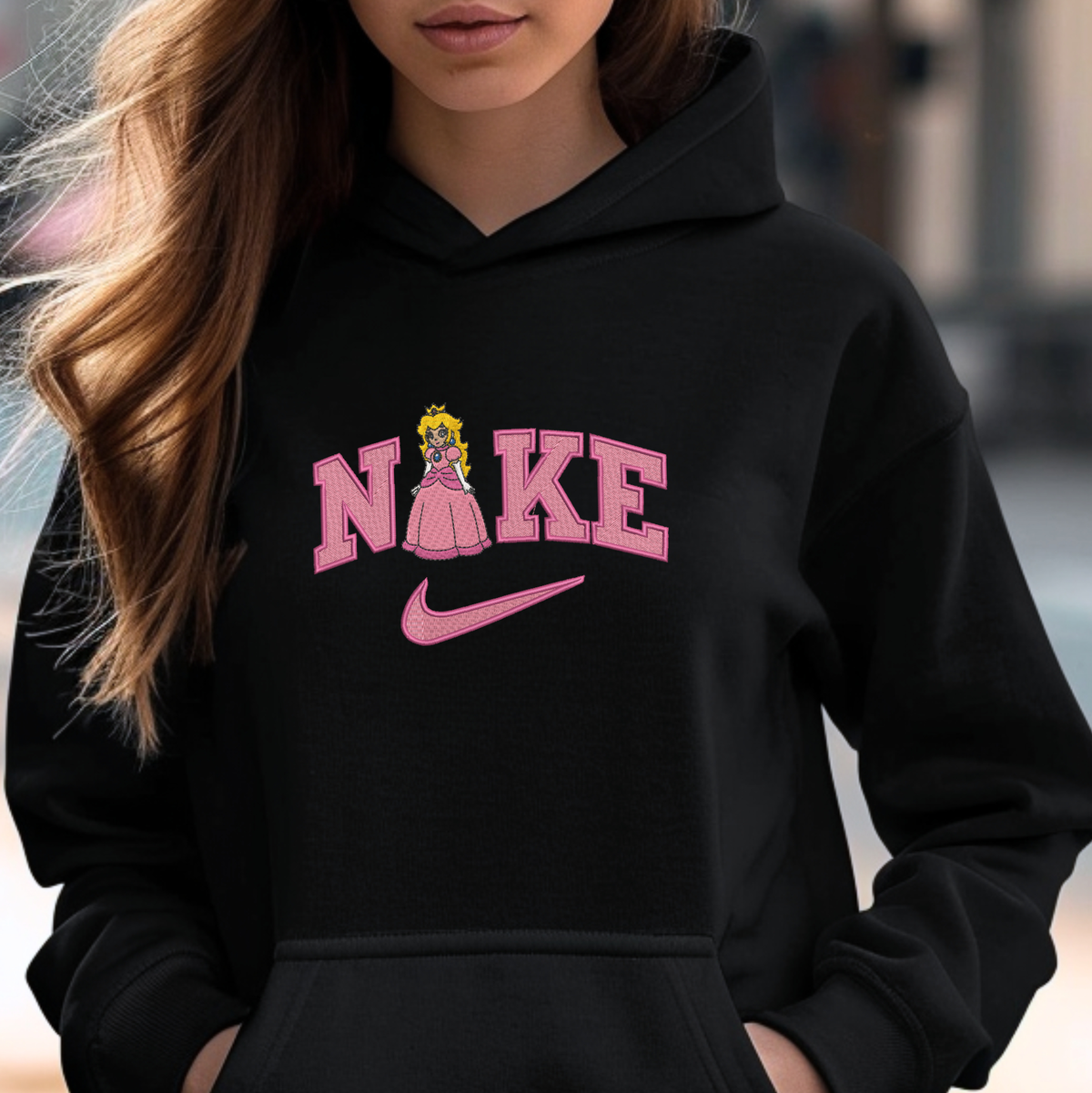 A woman wearing a black hoodie with a pink graphic that reads 'NIKE' styled like the Nike logo, featuring a cartoon princess peach inside the letter A and a pink swoosh underneath.