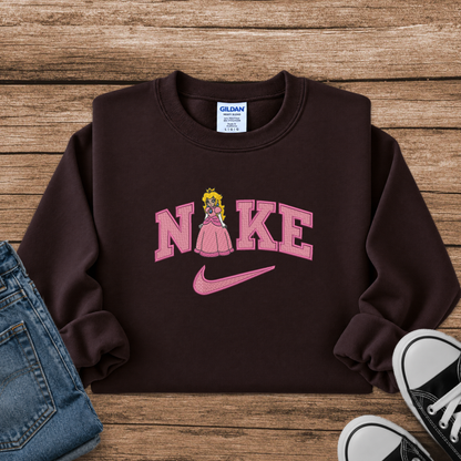 A dark brown crewneck sweatshirt featuring a pink 'Nike' graphic with a character resembling Princess Peach and a pink swoosh, styled in a flat lay on a wooden background with jeans and sneakers.