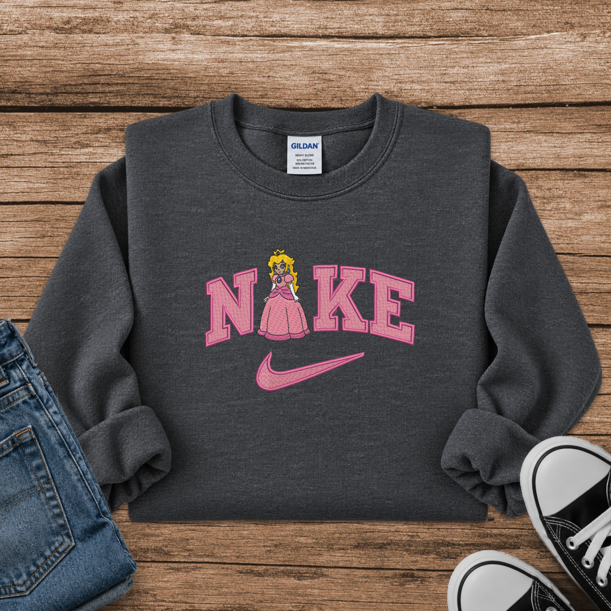 A dark gray crewneck sweatshirt styled in a flat lay on a wooden surface, featuring a pink 'NIKE' graphic with a princess character in the letter 'I' and a pink swoosh. Blue jeans and black sneakers are partially visible as styling props.