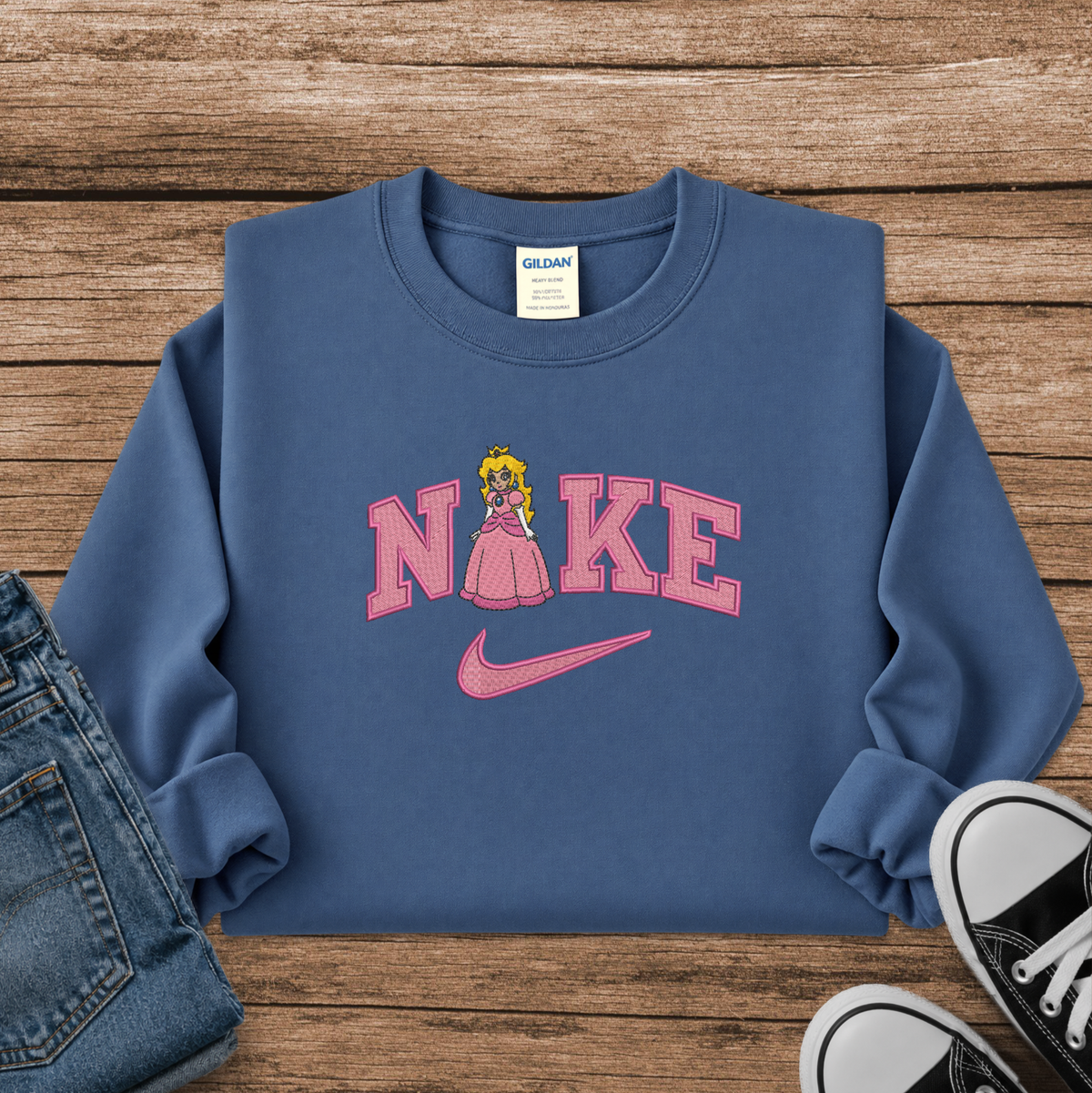 A blue crewneck sweatshirt styled in a flat lay on a wooden surface, featuring a pink 'Nike' graphic with a Princess Peach figure and a pink swoosh. Blue jeans and black sneakers are partially visible as styling props.
