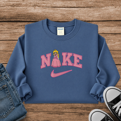 A blue crewneck sweatshirt styled in a flat lay on a wooden surface, featuring a pink 'Nike' graphic with a Princess Peach figure and a pink swoosh. Blue jeans and black sneakers are partially visible as styling props.