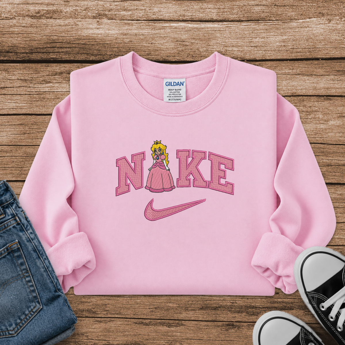A pink long-sleeve sweatshirt laid flat on a rustic wood background, featuring a 'Nike' wordmark with a princess peach character in a pink dress and a pink swoosh. A Gildan brand tag is visible inside the collar.